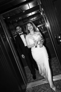 A bride in a white dress holding flowers and a groom in a tuxedo happily exit an elevator, both smiling and joyful—a perfect moment captured by a skilled photographer with a documentary approach to wedding photography.