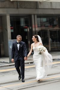 A bride in a white gown and a groom in a black tuxedo hold hands and smile at each other while walking across a city street, capturing an editorial photography moment full of romance and urban charm.