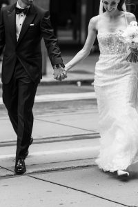 A bride in a white gown holds hands with a groom in a dark suit as they walk together outdoors. Captured by a talented photographer, the black and white editorial photo is cropped above their heads, focusing on their outfits and clasped hands.
