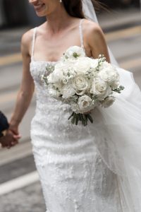 A bride in a white beaded wedding dress holds a bouquet of white roses and small flowers while standing outside, smiling, and holding hands with someone as the wedding photographer captures the joyful moment.