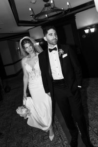 A bride in a white dress and veil holds a bouquet, standing next to a groom in a tuxedo. Both look away from the camera with serious expressions in a formal indoor setting. Captured in black and white, this wedding photograph conveys a timeless, documentary style.