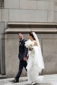 A bride in a white dress and veil holds a bouquet of white flowers while walking hand in hand with a groom in a dark suit, both smiling onto the scene against a stone building background, capturing an elegant wedding moment.