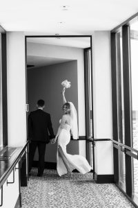 A bride in a wedding dress and veil smiles, holding a bouquet up high, while walking hand-in-hand with a groom in a suit down a hallway. Captured by a talented Toronto photographer, the black and white image exudes an elegant, editorial feel.