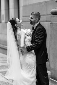 A bride and groom, both smiling, stand close together on a city sidewalk in this editorial-style image. The bride wears a long veil and a lace dress, while the groom is in a suit. Captured by an experienced wedding photographer, they look happy and joyful on their wedding day.