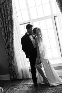 A bride and groom stand close together by a large window with curtains in this editorial black and white photograph. The bride holds a bouquet and wears a veil, while the groom is in a tuxedo. Captured by a Toronto photographer, the image exudes timeless elegance.