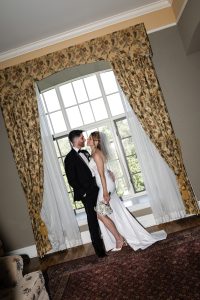 A bride and groom stand smiling, facing each other in front of a large window with patterned curtains. Captured in a documentary wedding photography style, the bride wears a white dress and veil; the groom is in a black suit.