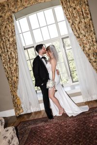 A bride and groom share a kiss in front of a large window with floral curtains, captured beautifully by their wedding photographer. The bride wears a veil and holds white flowers, while the groom wears a black tuxedo. This timeless moment reflects the artistry of wedding photography.