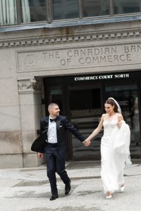A bride and groom, smiling and holding hands, walk onto the steps outside the Canadian Bank of Commerce building. The bride wears a white gown and veil; the groom wears a tuxedo, capturing an elegant wedding moment in an editorial style.