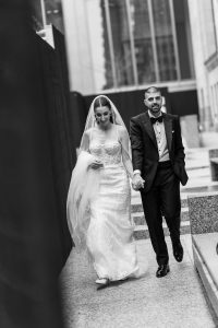 A bride and groom walk hand in hand outdoors, smiling, as they step onto stone steps framed by modern city buildings. The bride wears a white gown and veil, while the groom wears a tuxedo—captured beautifully in this moment of wedding photography.