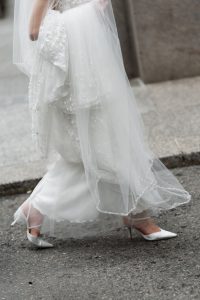 Close-up of a bride walking onto a city sidewalk, wearing a white wedding dress with a beaded veil and white high-heeled shoes. Her face is not visible in the image, giving the scene an elegant, editorial touch perfect for a modern wedding narrative.
