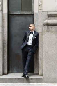 A man in a black tuxedo and bow tie stands leaning against a stone wall with his hands in his pockets, looking to the side as a photographer captures the moment, evoking a timeless wedding atmosphere.