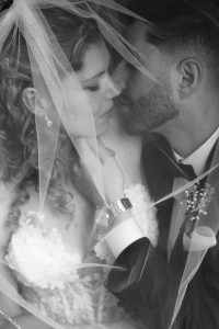 A bride and groom sit close under a veil, gazing lovingly at each other. The groom gently holds the bride’s chin as they almost kiss, dressed in wedding attire. Captured in black and white, this editorial photography image beautifully conveys a documentary feel of intimate romance.
