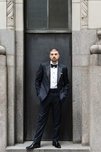 A man in a black tuxedo with a bowtie stands in a stone doorway, hands in his pockets, looking at the camera with a serious expression, evoking an editorial wedding portrait.