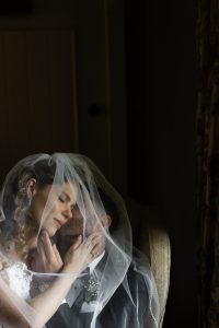A bride and groom sit closely, touching faces and embracing tenderly under a sheer veil—softly lit in a peaceful, intimate atmosphere that beautifully captures the essence of wedding photography. With the bride’s eyes closed, this moment feels like a fleeting scene from a romantic documentary.