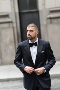 A man wearing a black tuxedo with a bow tie stands outdoors in front of a stone building, adjusting his jacket and looking to the side, as if posing for a wedding photographer.