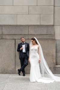 A bride in a white gown and veil stands beside a groom in a dark tuxedo, both with serious expressions, as their wedding photographer captures the moment against the backdrop of a stone wall.
