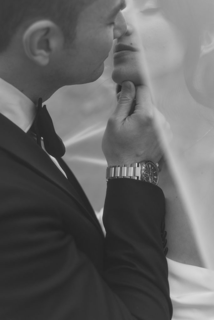 A groom in a suit gently lifts a bride’s chin for a kiss, both partially covered by a veil. Captured by a skilled Toronto photographer, the black and white image creates a romantic and intimate atmosphere.