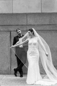 A bride in a lace gown and veil poses playfully for the photographer in front of a groom in a tuxedo, who leans against a stone wall, watching her with a relaxed expression.