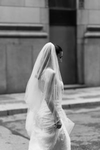 A bride in a white wedding dress and veil walks onto a city street, captured in black and white by the photographer for an editorial shoot.