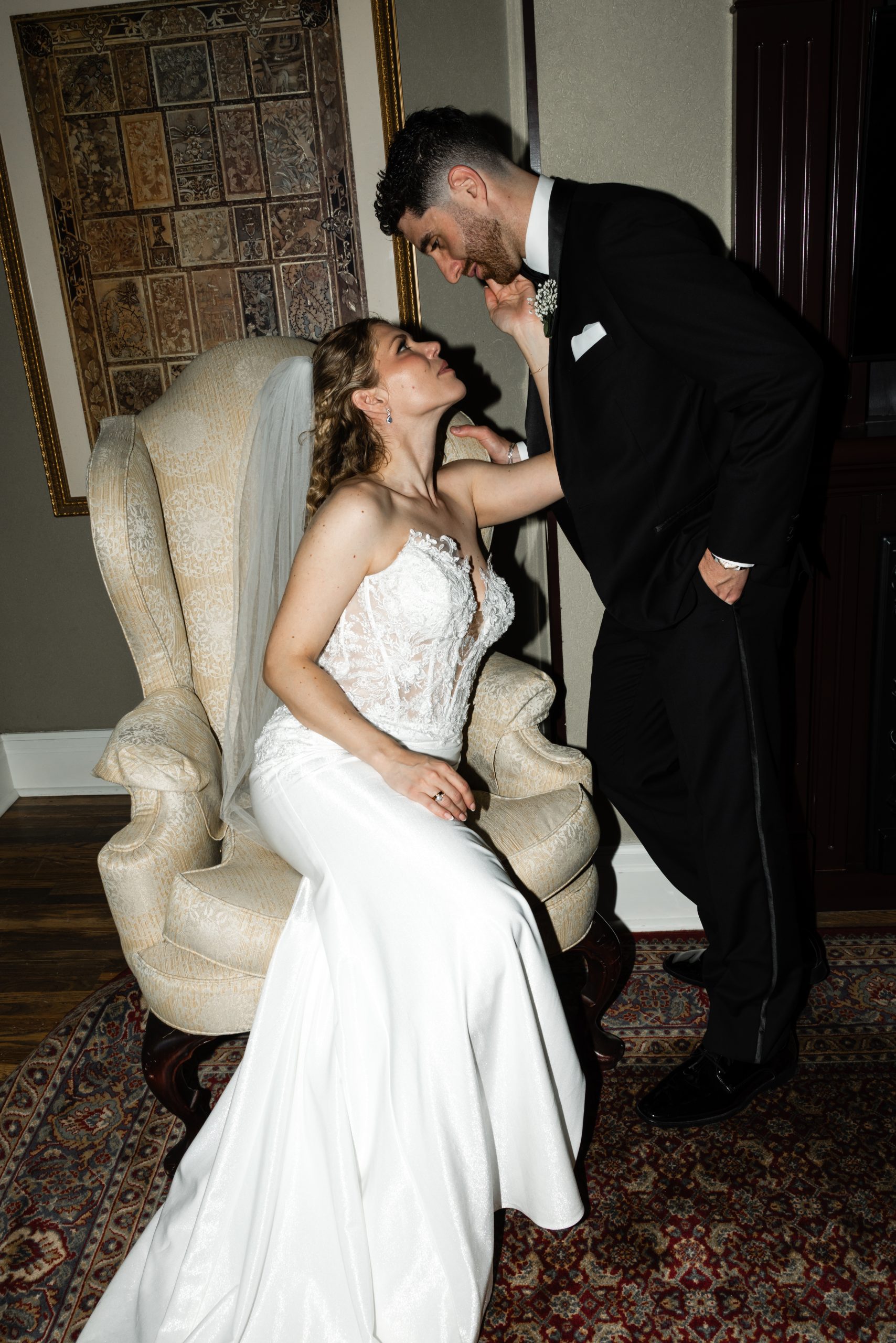 A bride in a white dress sits on a chair, gently touching the face of a groom in a black suit who leans toward her, both smiling lovingly—a tender moment captured in true editorial wedding style.