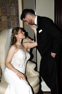 A bride in a white gown sits on a chair, gazing up and touching the face of a groom in a black suit who leans toward her, both sharing an affectionate moment captured in elegant wedding photography.