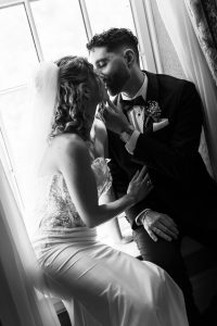 A bride and groom sit closely by a window, sharing an intimate kiss. The bride wears a veil and a strapless gown; the groom is in a suit with a boutonniere. This black and white wedding photography captures the timeless romance of their special day.