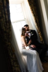 A bride and groom sit closely together by a sunlit window, sharing an intimate moment. The image is soft-focused, creating a dreamy and romantic atmosphere with draped curtains framing the couple—an exquisite example of editorial photography that captures the essence of their love.
