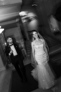 A bride in a wedding dress smiles at the camera while a man in a tuxedo stands nearby. Captured by an editorial photographer, the black and white photo uses motion blur to create a dreamy, surreal effect.