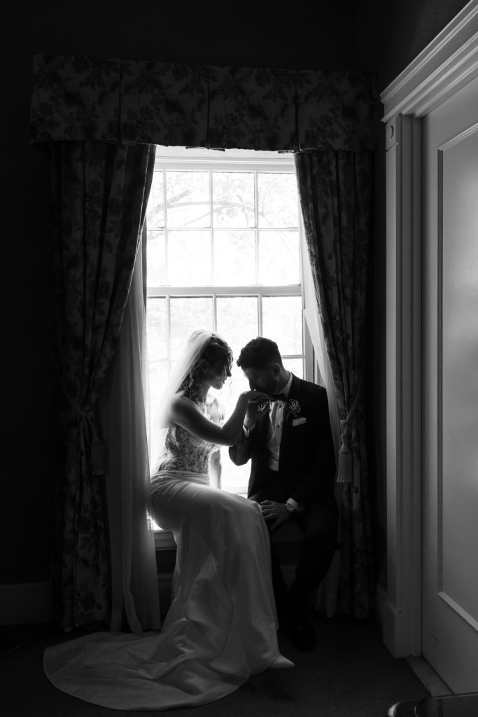 A bride and groom sit together by a window, the groom kissing the bride’s hand. Soft light from the window highlights them, creating a romantic black-and-white scene reminiscent of a Toronto editorial wedding.