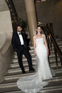 A bride in a white gown and veil stands beside a groom in a black tuxedo on a marble staircase with gold railings, both looking in different directions, as if captured by an editorial photographer.