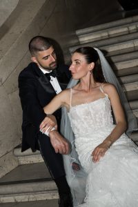 A bride in a white wedding dress and veil sits on stairs, holding hands and looking onto a groom in a black tuxedo who is seated beside her. Both appear relaxed and content, creating an editorial feel to the moment.