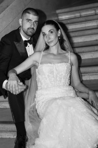 A bride in a beaded gown and veil sits on stairs, holding hands with the groom in a black tuxedo. Both look at the camera, posing together in an elegant, editorial style that highlights refined wedding photography.