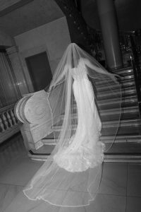 A bride in a long, elegant gown and flowing veil stands on a marble staircase, facing away from the camera. The wedding scene is captured in black and white photography.