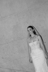 A woman in a beaded, strappy wedding dress and long veil stands against a stone wall, looking to the side. Captured by an editorial photographer, the black and white photo highlights her elegant pose as light falls gently onto her profile.
