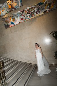 A bride in a white gown and veil stands on marble stairs beneath a colorful, abstract sculpture attached to a beige stone wall, captured in striking editorial photography.