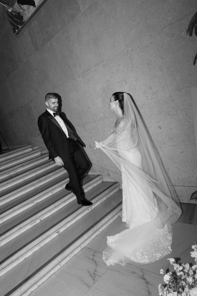 A groom in a tuxedo descends stairs, smiling at a bride in a white dress and veil who stands at the bottom, holding her gown. Captured in black and white by a skilled wedding photographer, the scene is elegant and formal with an editorial quality.
