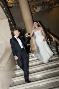 A bride in a white gown and veil holds hands with a groom in a dark tuxedo as they walk down elegant stairs, both smiling—a timeless moment perfectly captured by the photographer’s editorial style.