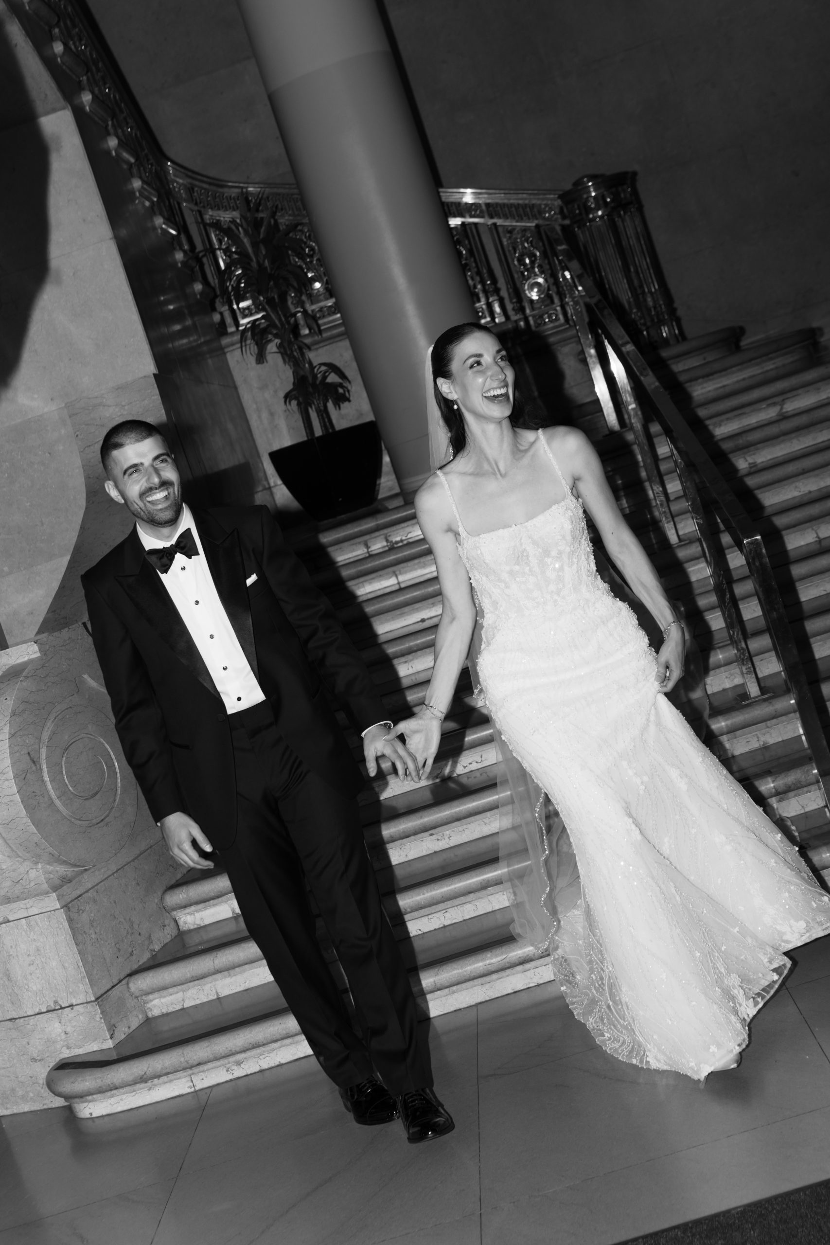 A smiling couple in formal wedding attire holds hands and walks onto ornate marble stairs, captured in elegant black and white editorial photography.