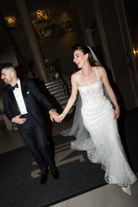 A bride in a white gown and veil laughs while holding hands with a groom in a black tuxedo as they walk indoors, possibly at their wedding reception, smiling brightly onto the photographer capturing their joyful moment.