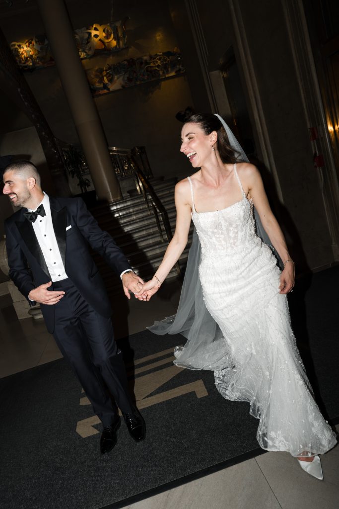 A bride in a white gown and veil laughs while holding hands with a groom in a black tuxedo as they walk indoors, possibly at their wedding reception, smiling brightly onto the photographer capturing their joyful moment.