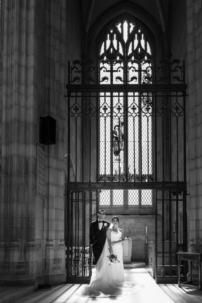 A bride and groom stand together in a grand church during their Toronto wedding, sunlight streaming through tall, ornate windows behind them, creating dramatic shadows on the floor.