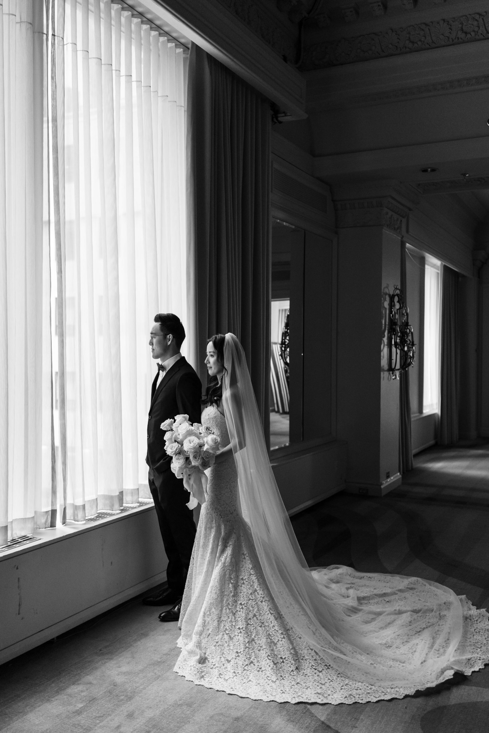 A bride and groom stand side by side by a large window, gazing outside as if posing for an editorial. The bride holds a bouquet of flowers and wears a long lace gown with a train, her elegance perfectly captured by the photographer. The room is elegant with tall curtains and ornate details.
