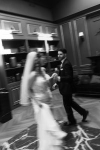 A black-and-white, blurred photo in a documentary style shows a bride and groom dancing indoors, holding hands. The room has bookshelves, paneling, and a couch in the background, creating an elegant and intimate atmosphere—an authentic moment captured through Toronto photography.