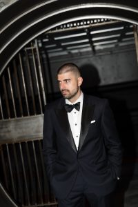 A man in a black tuxedo stands with hands in pockets in front of a large circular vault door, looking off to the side, creating an editorial mood reminiscent of a high-fashion wedding shoot.