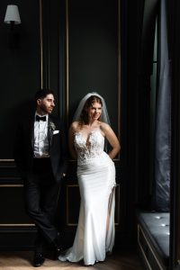 A bride in a white dress stands smiling by a window next to a groom in a black tuxedo, who is leaning casually against a dark wall, creating an editorial moment skillfully captured by the photographer in Toronto.