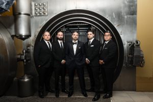 Five men in black suits and bow ties stand in front of an open, round vault door, posing formally and looking at the camera in a well-lit setting, as the photographer captures the moment with precision.