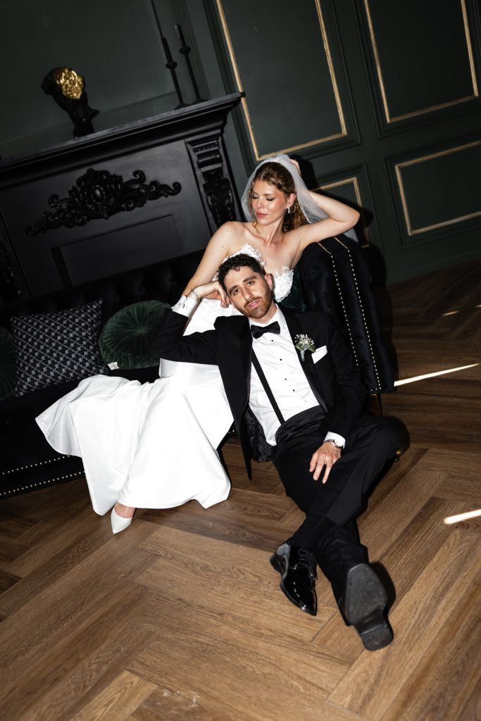 A bride in a white dress sits on a couch, smiling, while the groom in a black tuxedo sits relaxed on the floor in front of her. Captured in an elegant, dark-paneled room, their playful and happy demeanor reflects the candid spirit of a documentary wedding in Toronto.