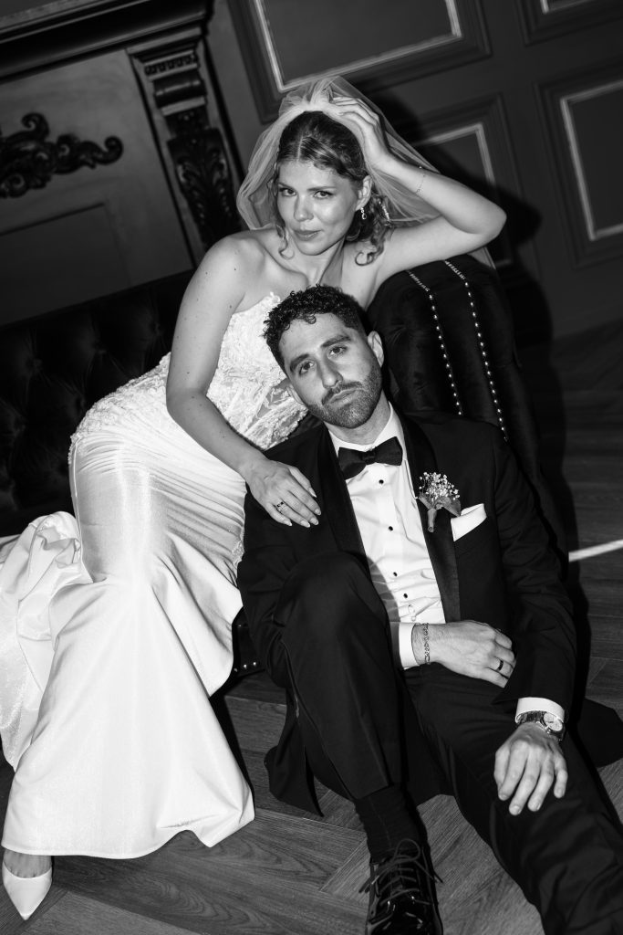 A bride in a white gown and veil leans on a sofa while a groom in a tuxedo sits on the floor in front of her. Both have relaxed expressions, captured in an elegant black and white editorial style that perfectly encapsulates the charm of a Toronto wedding.