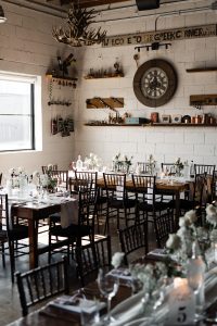 A sunlit rustic dining room with wooden tables, dark chairs, white flowers, and glassware—perfect for a wedding celebration. Shelves with decor and a large clock hang on a white brick wall, while antlers and bottles add charm. This intimate space in Toronto offers endless inspiration for any photographer.