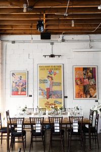 A rustic dining table with floral centerpieces is set for eight beneath vintage movie posters on a white brick wall, evoking the atmosphere of an editorial photoshoot in an industrial-style room with exposed wooden beams and hanging lights.